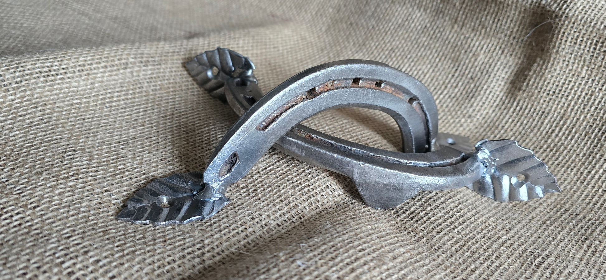 A pair of silver-colored metal horseshoe cupboard handles with leaf detailing, placed on a burlap fabric surface.
